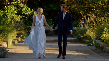 Newlyweds walking in the park. Action.A beautiful blonde bride in a white dress with heels together with her bearded groom in a black suit holding hands smiling at each other.