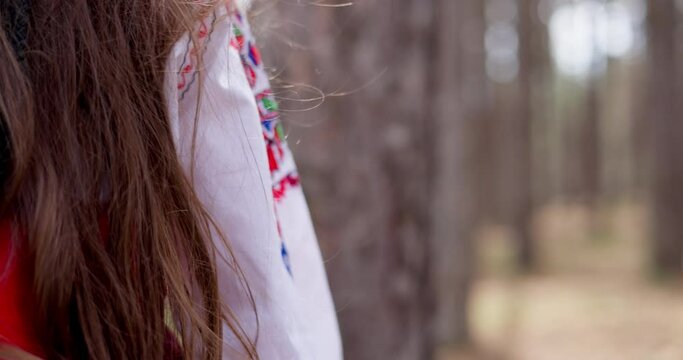 Bulgarian girl in traditional folklore costume red white dress with shevitsa touching with hand trees in forest of majestic mountain, 4k slow motion video. Bulgaria nature.