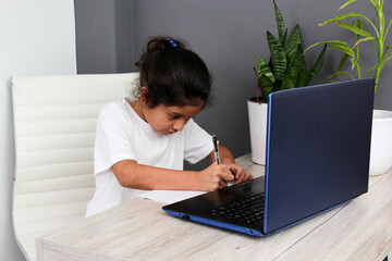 9-year-old Latino girl with glasses does home schooling takes online classes at home on a desk with a laptop, studies, is surprised and participates in class