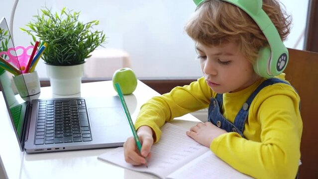 Cute smart caucasian primary School Child with headphones using laptop. Online tutor teaching digital class, web lesson on computer at home. Virtual education. Schoolboy girl study write do homework