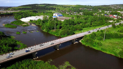 Obraz premium Aerial view of a river and a bridge with driving cars on a summer day. Clip. Summer village surrounded by green vegetation.