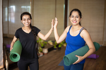 Two Beautiful young happy indian girls standing and holding yoga mat together after finishing her...