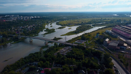 Aerial view of the city parts connected by the bridge. Clip. Wide river flowing along green vegetation and buildings. - obrazy, fototapety, plakaty