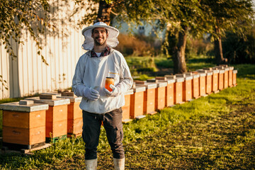 Young experienced beekeeper in protective workwear working at apiary and feeding hives. He is happy and holding a jar of honey while looking at the camera and smiling. - obrazy, fototapety, plakaty