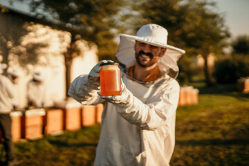 Portrait of a handsome beekeeper in a protective uniform standing with honey, tasting fresh...