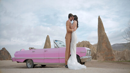Beautiful bride and groom in wedding dress and suit near pink vintage cabriolet car. Action....