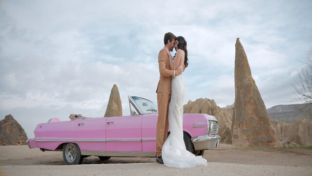 Beautiful bride and groom in wedding dress and suit near pink vintage cabriolet car. Action. Wedding couple standing near a convertible on a natural background.
