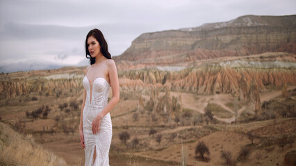 Woman wearing bride dress stands on the canyon landscape background. Action. Beautiful young brunette woman in front of picturesque valley.