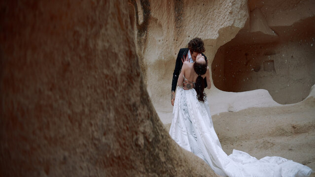 Stylish beautiful bride and groom under falling snow near rocks or caves. Action. Wedding video shooting in cappadocia.