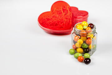 A red heart-shaped translucent gift box and a glass jar with colorful chocolate balls. Confess your love with all your heart