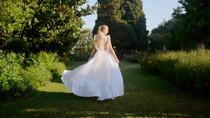 Beautiful young bride in a sunny summer park. Action. Woman in white dress in an alley with small bushes.