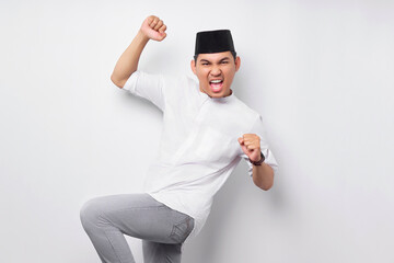 Excited young Asian Muslim man raising fists and celebrating victory on Ramadan isolated over white...