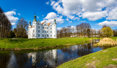 Fototapeta na wymiar Panoramic view of the majestic Schloss Ahrensburg castle, a white Renaissance castle surrounded by a moat, reflecting its grandeur amidst the castle park in April, with blue skies and sunlight.