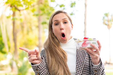 Young blonde woman holding a three dimensional puzzle cube at outdoors surprised and pointing side