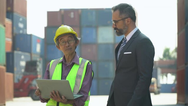senior asian engineer holding laptop computer talking with business man in suit checking list inventory loading Containers box at warehouse logistic in Cargo . owner and worker diversity