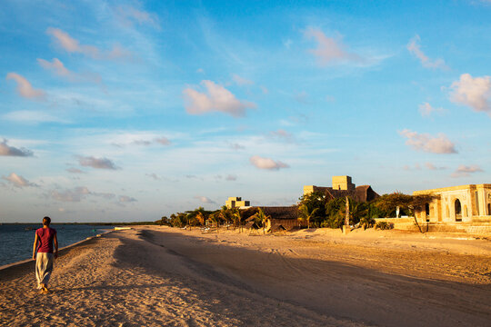 LAMU, INDIAN OCEAN, KENYA, AFRICA. A young woman walks down the empty white beach of Manda Island under bright blue sky.
