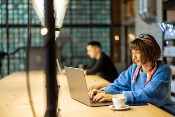 Young stylish woman works on laptop while sitting by shared table with a coffee drink at modern cafe. Concept of remote work from public place, digital freelance and modern lifestyle