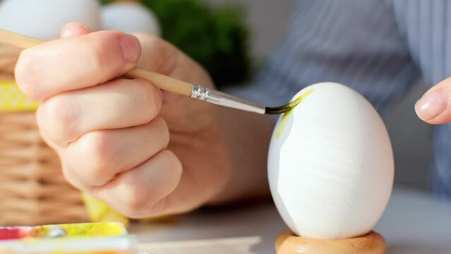 Hand of a young woman decorating a chicken egg with a brush and paints, close-up. Decoration for the spring holiday. Preparation for the celebration of Happy Easter day.