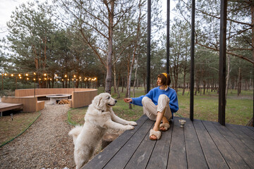 Young woman cares her dog while sitting on a porch of wooden house in pine forest, spending leisure time together and resting at countryside