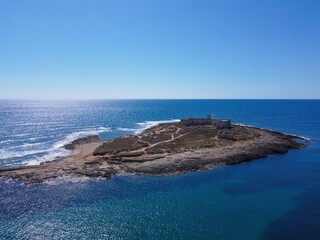 Aerial drone view of the island and beach of Isola delle Correnti with and old lighthouse surrounded by clear turquoise sea water. Southernmost point in Sicily, Portopalo di Capo Passero, Italy.