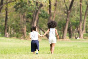 African and Caucasian little cute girls holding hands, walking together at green park, wear white vest. Diversity children friends have fun playing summer outdoors