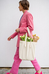 Stylish woman in pink suit walks with eco handbag full of flowers on white background. Canvas bag...
