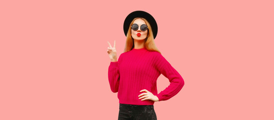 Portrait of stylish young woman model blowing her red lips sends kiss wearing pink knitted sweater and black round hat isolated on background