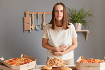 Image of sad ill sick woman with brown hair wearing white T-shirt sitting at table eating junk food and having stomachache, has food poisoning, touching her belly.