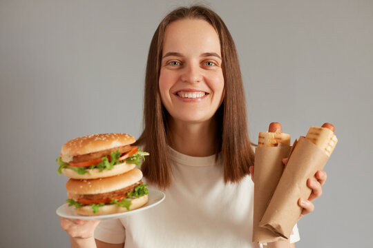 Indoor shot of smiling cheerful young adult woman with dark hair dressed in white T-shirt holding out big burger sandwich and sausages in dough, giving you fast food snack.