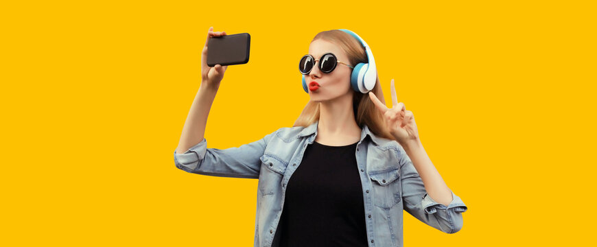 Portrait of stylish young woman taking selfie with smartphone in wireless headphones listening to music and blowing her lips sends kiss isolated on yellow background