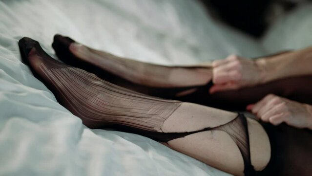 woman is tearing her nylon pantyhose sitting on bed with white linen, closeup of legs, foot fetish