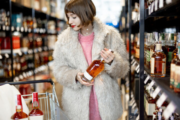 Woman chooses strong alcohol standing with shopping cart between rows of bottles in a supermarket....