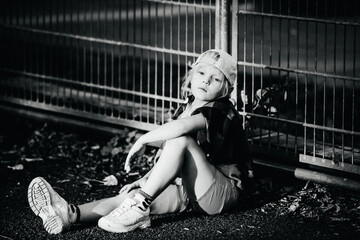 Obraz premium Black and white portrait of a stylish blonde model child in casual clothes and a cap sits on the ground leaning against a fence