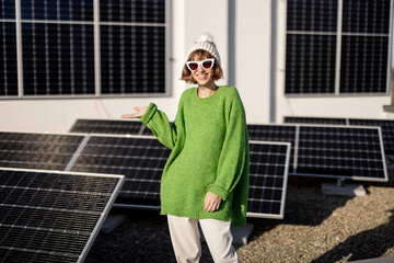 Woman in green sweater and winter hat stands on a rooftop of her house with a solar power plant...