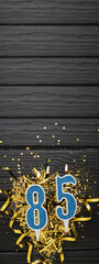 Number 85 blue celebration candle and gold confetti on dark wooden background. 85th birthday card....