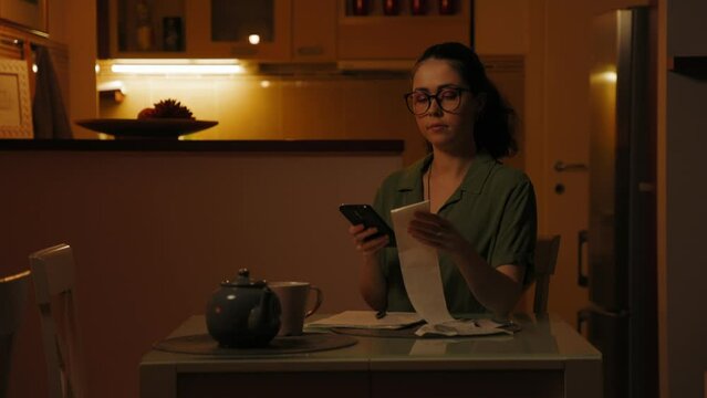 Young Caucasian woman wearing glasses is sitting at table and using smartphone, holding paper receipt in her hand. Domestic accounting. Concept of financial applications and tax return.