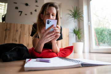 Fototapeta premium Casual young woman, a student, lying on a living room floor browsing on her mobile phone