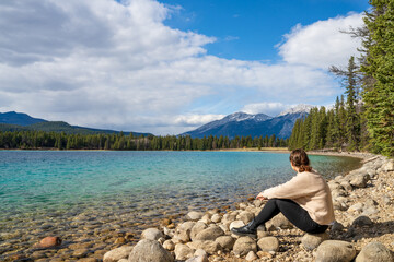 Fototapeta na wymiar Young girl sitting by the lake shore enjoying the nature scenery. Summer time in Lake Annette, Jasper National Park, Canadian Rockies, Alberta, Canada.