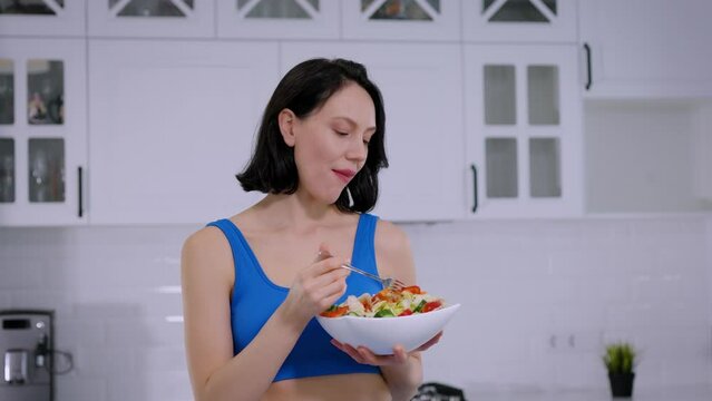 A middle-aged brunette woman eats salad in a white kitchen with gusto