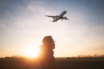 Man with backpack looking up to airplane landing at airport during beautiful sunset. ..