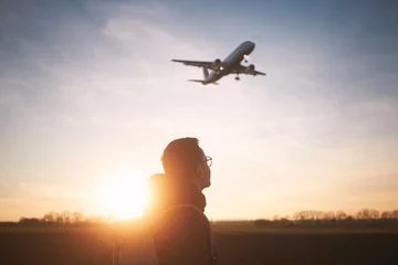 Fotobehang Man with backpack looking up to airplane landing at airport during beautiful sunset. .. © Chalabala