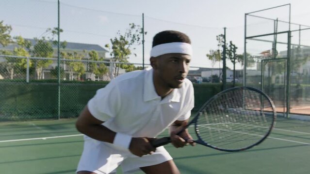 Athletic young black man in headband hitting ball with tennis racquet while playing at sports court