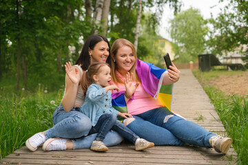 A young happy cheerful LGBT married couple with a funny laughing daughter communicate with their family via video communication on a smartphone. People wave at the camera in greeting