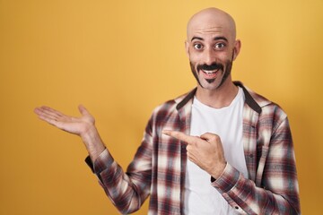 Hispanic man with beard standing over yellow background amazed and smiling to the camera while presenting with hand and pointing with finger.