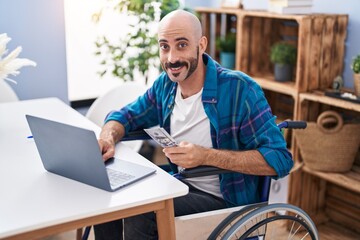 Young hispanic man using laptop holdig dollars sitting on wheelchair at home