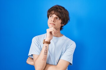 Hispanic young man standing over blue background with hand on chin thinking about question, pensive expression. smiling with thoughtful face. doubt concept.