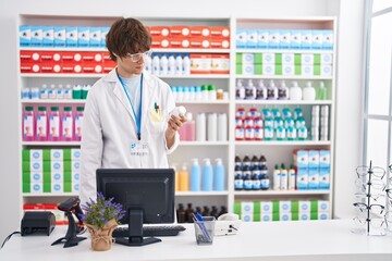 Young blond man pharmacist using computer holding pills bottle at pharmacy