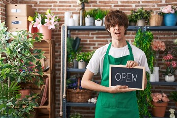 Hispanic young man working at florist holding open sign clueless and confused expression. doubt concept.