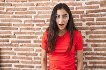Young teenager girl standing over bricks wall afraid and shocked with surprise and amazed expression, fear and excited face.