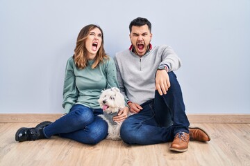 Obraz na płótnie Canvas Young hispanic couple sitting on the floor with dog angry and mad screaming frustrated and furious, shouting with anger. rage and aggressive concept.